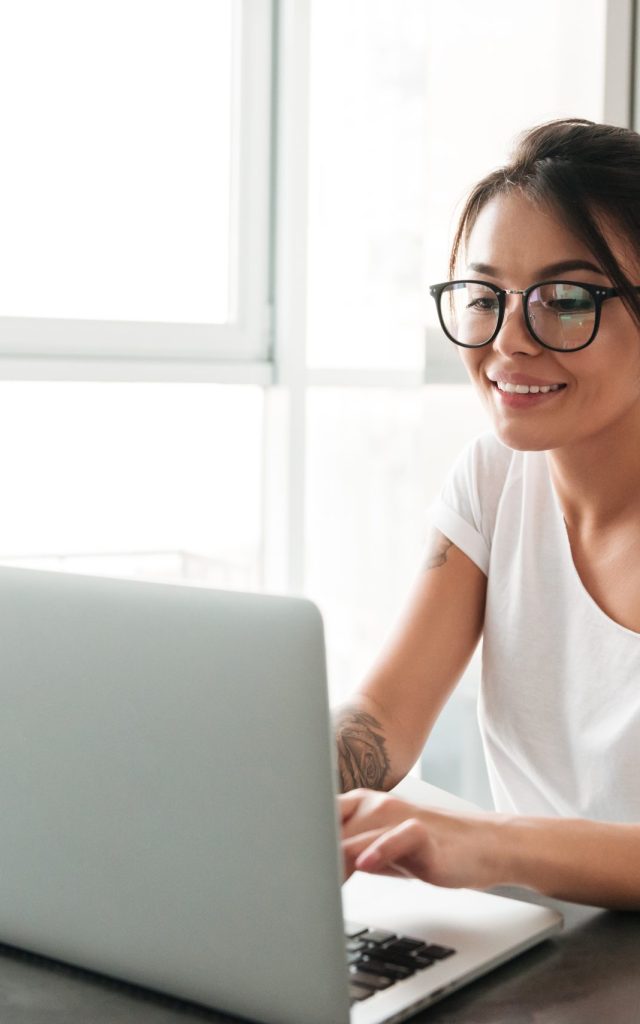 Image of happy young woman sitting at the kitchen in home. Looking aside chatting by laptop computer.